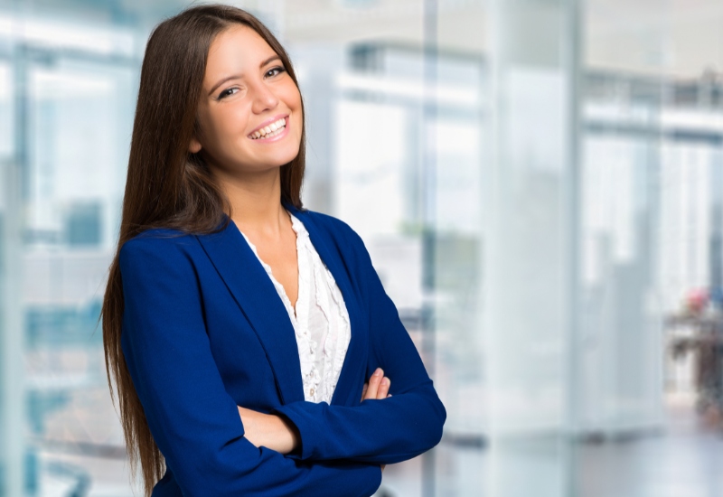 A professional woman with long brown hair smiles confidently while wearing a blue blazer over a white blouse. She stands in an office environment with a blurred background of glass windows.