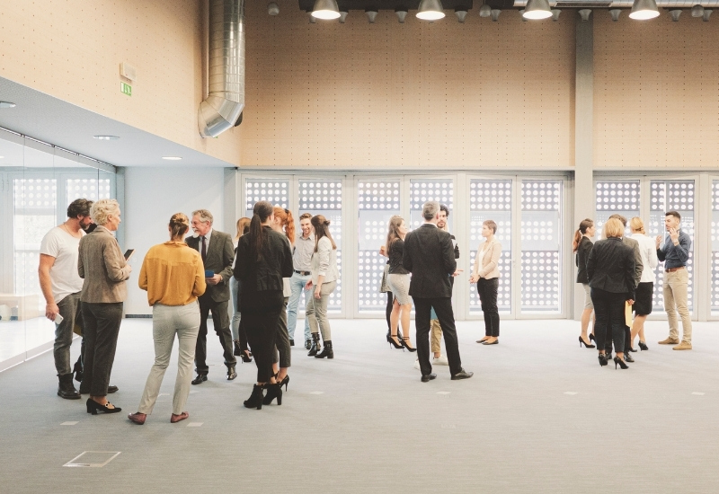 A group of people engaging in conversation at a networking event in a spacious, modern setting. The attendees are dressed in business attire, with a mix of men and women gathered in small groups.