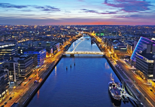 An aerial view of a cityscape during twilight, featuring a river with a lit bridge and buildings along the banks. The sky shows a gradient of colors from yellow to dark blue as city lights begin to illuminate the streets.
