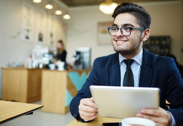 A young man in business attire, wearing glasses, sits at a table holding a tablet in a café. He is smiling while looking at the device, with a coffee cup in front of him and a coffee counter visible in the background.