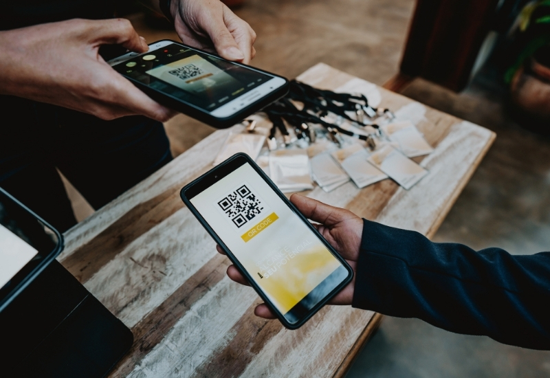 A person holding a smartphone displaying a QR code, while another person uses a device to scan it. In the background, a table with lanyards and identification tags is visible.