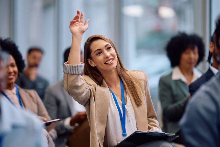 A woman with long hair raising her hand during a meeting, smiling. She is seated among a diverse group of people in a professional setting, wearing a blazer and a blue lanyard.