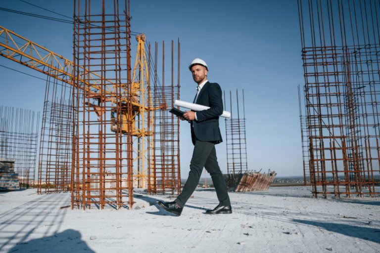 A man in a suit and hard hat walking on a construction site, holding blueprints. Steel rebar structures and a crane are visible in the background under a clear blue sky.