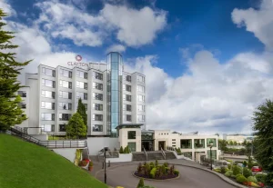 The exterior of the Clayton Hotel, featuring a modern multi-storey building, large windows, and a glass entrance. Lush green landscaping and a blue sky with scattered clouds are visible in the background.