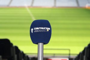 A microphone on a stand with a blue cover, featuring text that reads "Construction Job Expo," in front of a green sports field.
