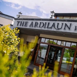 The entrance of The Ardilaun Hotel, featuring a large sign above the doors, surrounded by greenery and a bright sky in the background.