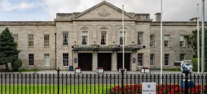 The front facade of a stone building featuring a portico with columns, large windows, and an emblem at the top. Green lawns and flower beds are in front, with a registration sign and flagpoles beside the entrance.