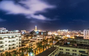 A nighttime cityscape view showing palms lining a street, with illuminated buildings and a lighthouse in the distance under a starry sky.